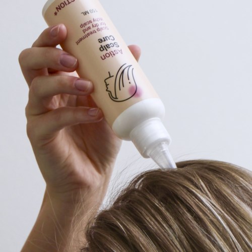 A person applies a scalp care shampoo to hair, holding a bottle with a label. The background is plain and white.