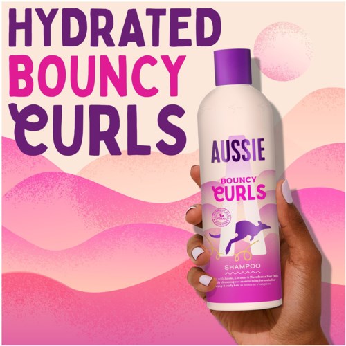 A hand holds a bottle of Aussie Bouncy Curls shampoo with purple cap, set against pink and peach wavy background.
