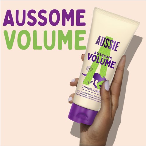 A hand holds a shampoo bottle with purple and green text, showing the product name, 'Aussome Volume Conditioner,' on a beige background.