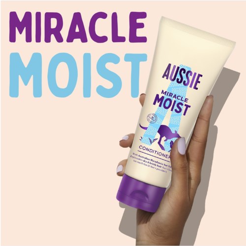 A hand holding a tube of Aussie Miracle Moist conditioner with a giraffe and elephant illustration on the tube.
