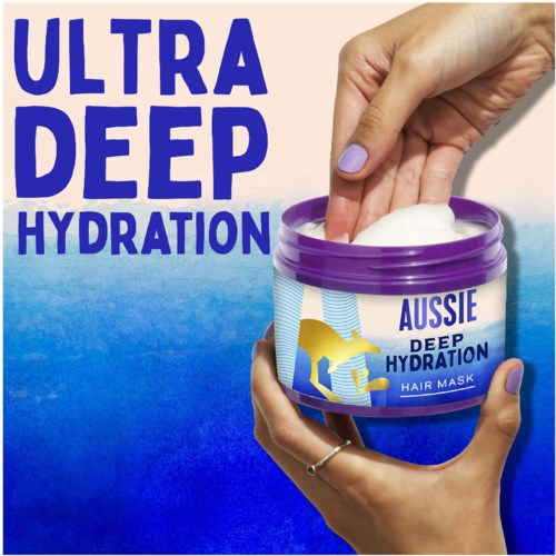 A hand is putting a white cream into a purple container labeled 'Aussie Deep Hydration Hair Mask' with a blue background.