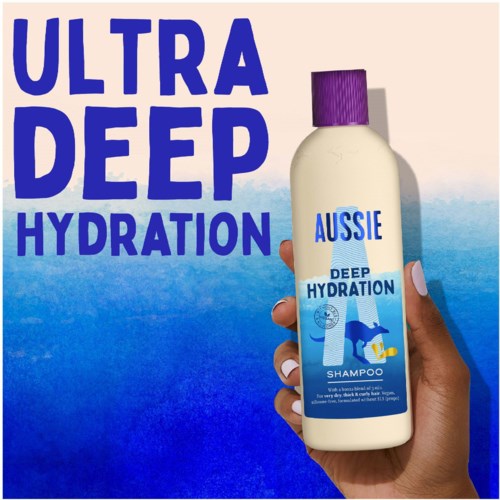 A hand holds a bottle of Aussie Deep Hydration shampoo, with bold blue text and a purple cap.