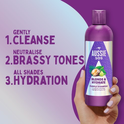 A purple shampoo bottle labeled 'Aussie SOS Blonde & Hydrate,' with a hand holding it, showing benefits for hair.