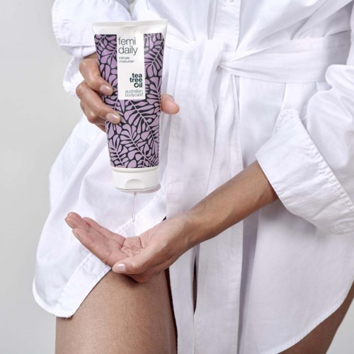 A woman in a white robe uses a tube of Femí daily tea tree oil on her skin.