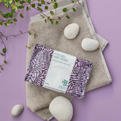 A pack of intimate wet wipes placed on beige towels, with three white stones and a green plant branch on a purple background.