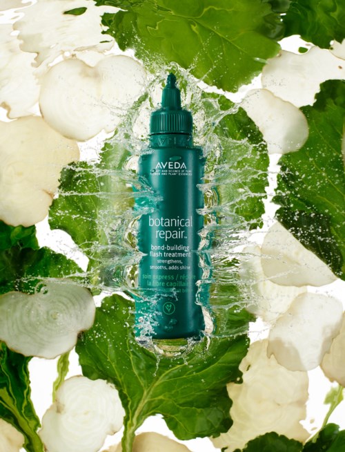 Eine blaue Flasche Aveda Botanical Repair Haartreatment, umgeben von grünen Blättern und weißen Blumen.