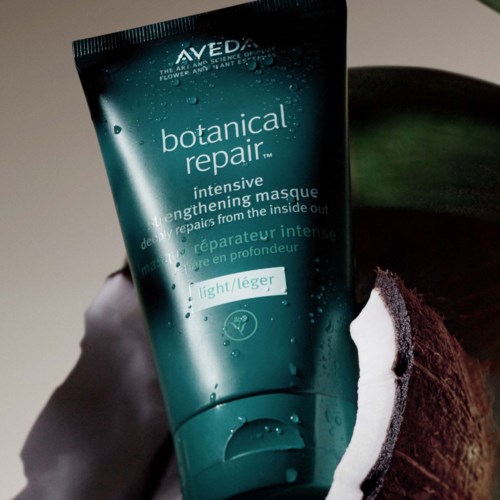 Ein tealfarbener Tube von Aveda botanical repair Haarkur mit Wassertropfen, gehalten von einer Hand vor dunklem Hintergrund.