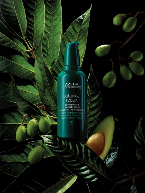 Eine blaue Flasche des Aveda botanical repair Haarprodukts umgeben von grünen Blättern und Früchten auf schwarzem Hintergrund.