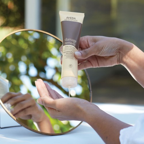 A person applies Aveda damage remedy hair treatment in front of a mirror outdoors.