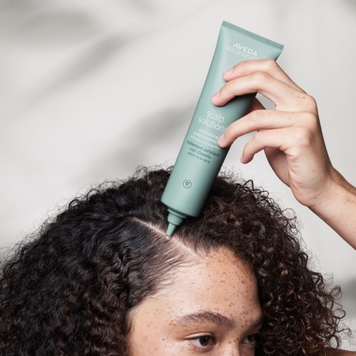 Eine Person trägt Haarbehandlung auf lockiges Haar mit einer Tube namens Aveda scalp solution auf.