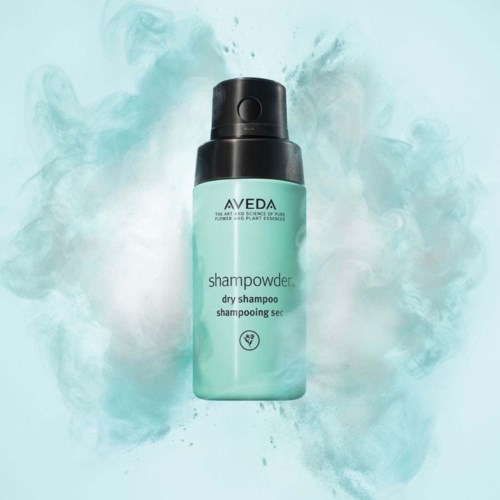 A bottle of Aveda dry shampoo with black spray cap, surrounded by light green powder or spray.