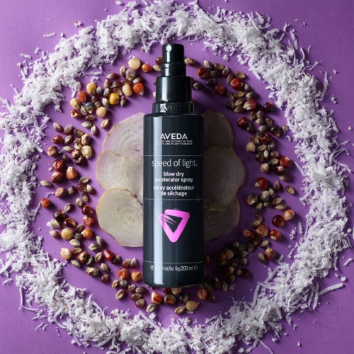 En svart sprayflaske merket 'Aveda' omgitt av frø, skall og hvitt strimlet materiale på lilla bakgrunn.