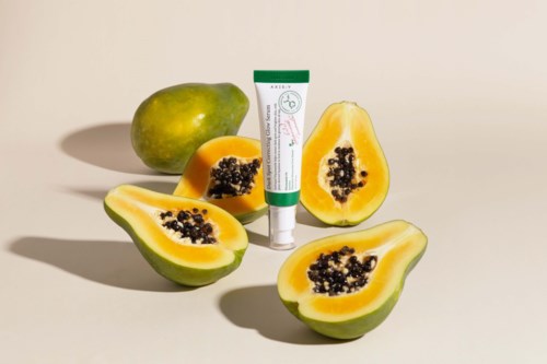 Drie helften van papaya en één hele papaya naast een tube huidverzorgingsproduct op een neutrale achtergrond.