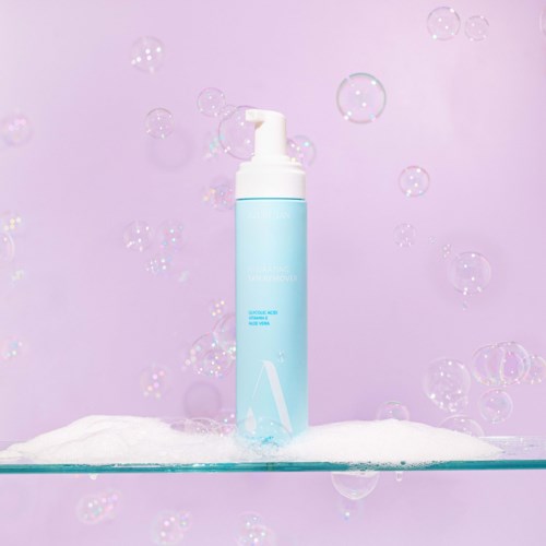Eine blaue Flasche mit Pumpe steht auf einer flachen Oberfläche, umgeben von Schaum und schwebenden Blasen vor einem pinken Hintergrund.