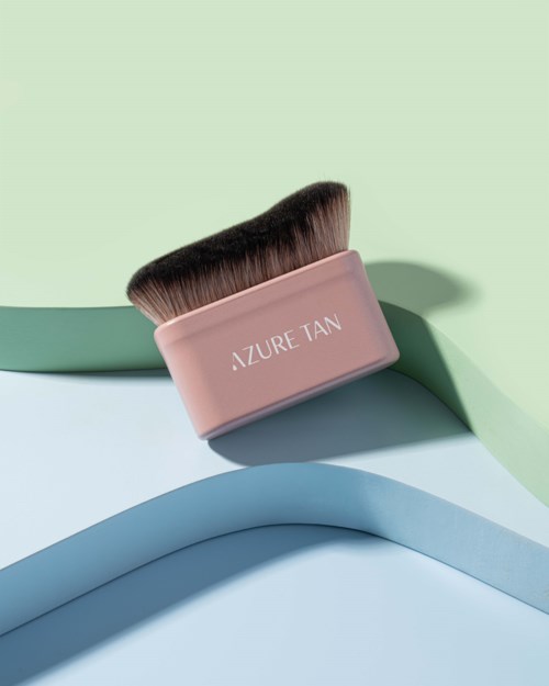 Eine Make-up-Bürste mit pinkem Griff, beschriftet 'AZURE TAN', liegt auf bunten Papierlagen.