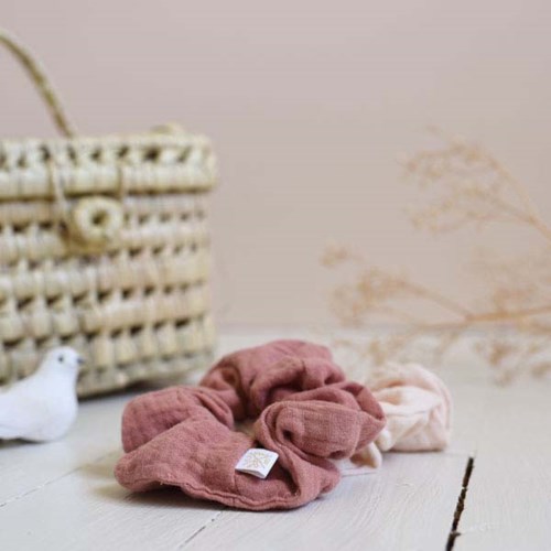 Soft fabric scrunchies in pink and beige rest on a white surface with a wicker basket and decorative plant in background.
