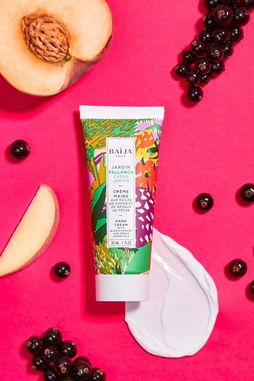 A colorful tube of hand cream surrounded by peach slices, blackcurrants, and a dollop of cream on a pink background.