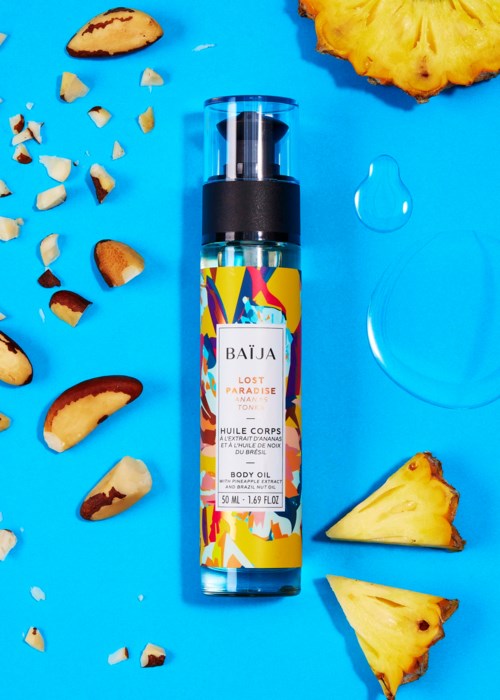 A body oil bottle labeled 'Lost Paradise' surrounded by pineapple pieces and crushed nuts on a blue background.