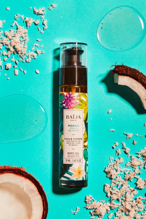 A bottle of body oil labeled Moana, surrounded by coconut pieces and small white grains on a blue background.