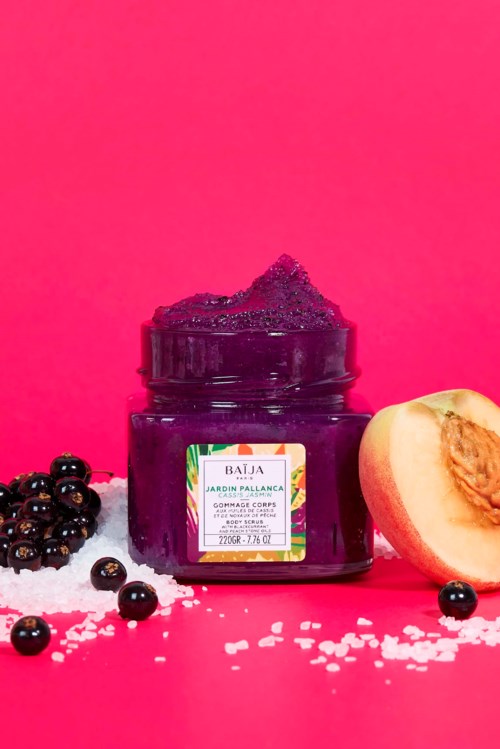 Dark purple body scrub in a jar with blackberries and peach slices on a pink background.