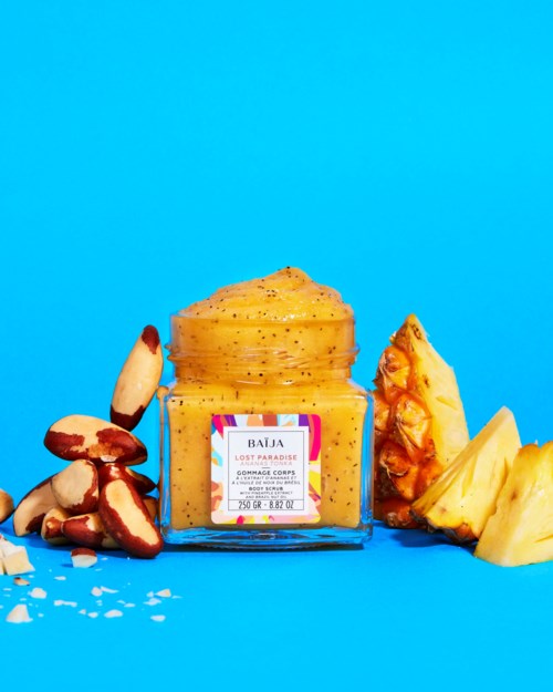 A yellow body scrub jar with tropical fruits around it on a blue background.