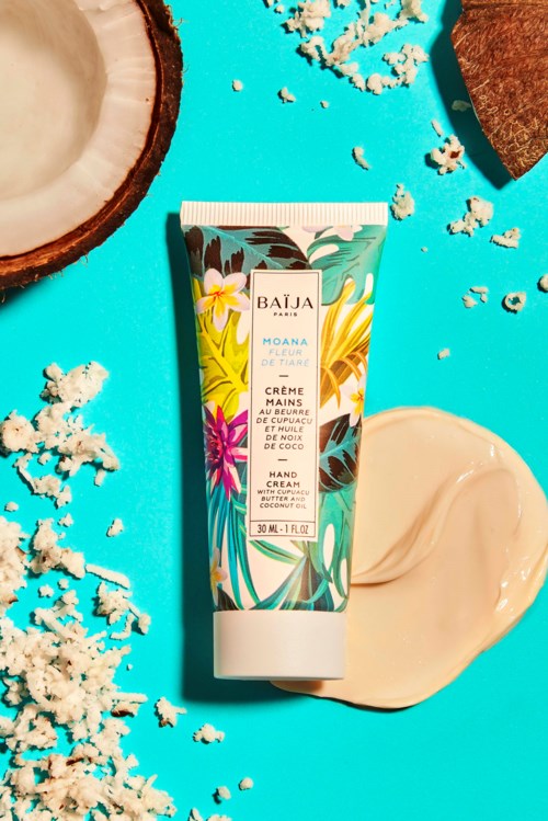 A tropical-themed hand cream tube with yellow, pink, and green leaves on a blue background, surrounded by coconut pieces.