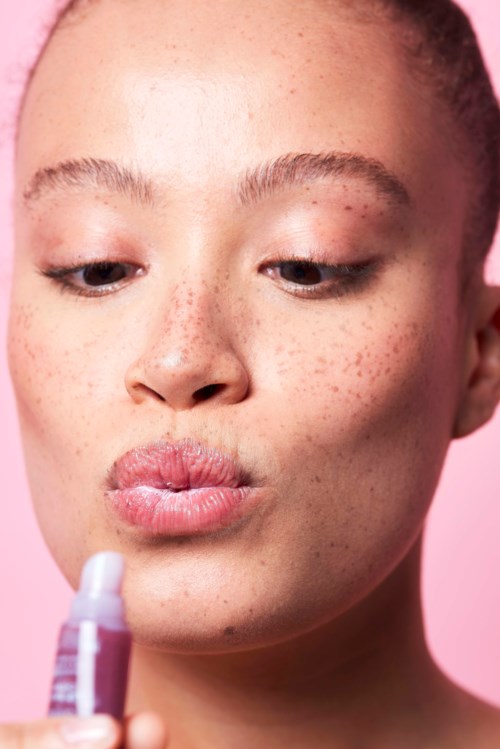 A woman with freckles looks at a small bottle, possibly a makeup or skincare product, against a pink background.