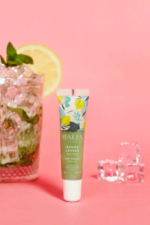 A tube of lip balm stands on a pink surface, with a mint and lemon drink in a glass and ice cubes nearby.