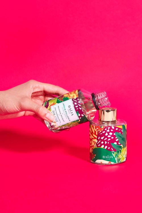A hand pours pink glitter from a colorful bottle into a matching container on a pink background.