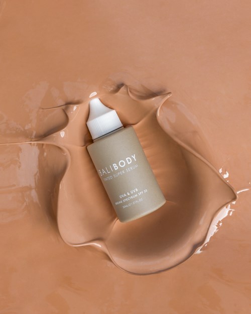 Een fles met het label 'BALIBODY' omgeven door gesmolten beige crème op een vlakke ondergrond.