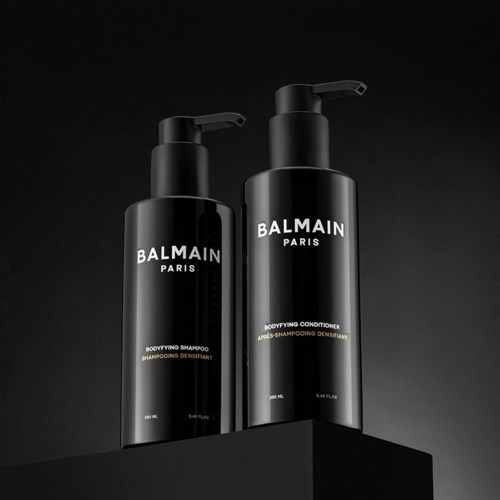 Två svarta flaskor med Balmain Paris kroppsshampoo och balsam på en svart yta. Minimalistisk design, vit och guld text.