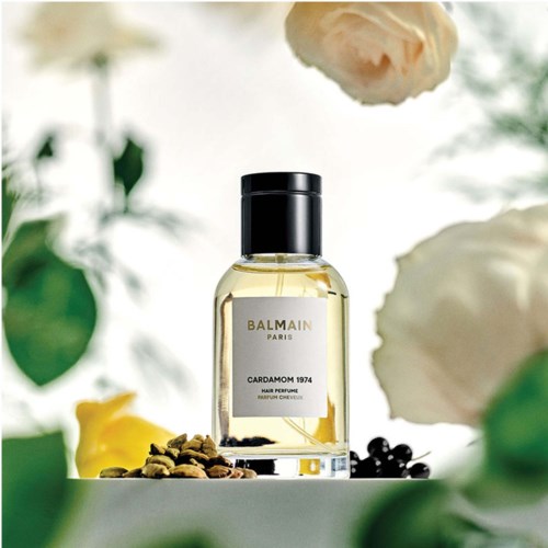 En parfymeflaske merket 'Balmain Paris Cardamom 1974' omgitt av blomster og krydder på en hvit overflate.