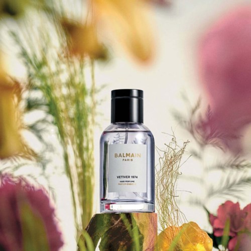 En tydlig parfymflaska märkt 'Balmain Paris Vetiver 1974' står bland rosa- och gulblommor och gröna växter.
