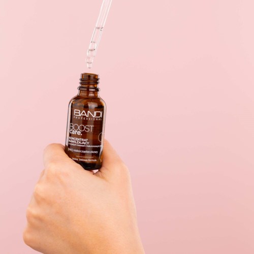 A hand holds a small brown bottle with a dropper, containing a liquid. The background is pink.