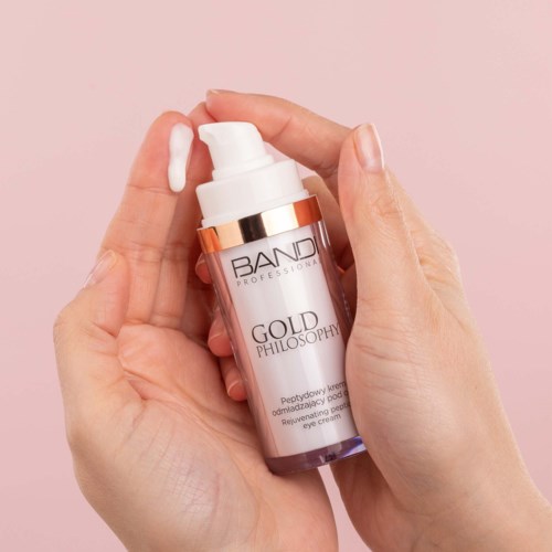 A hand holding a small bottle of eye cream with a pump, labeled "GOLD PHILOSOPHY."
