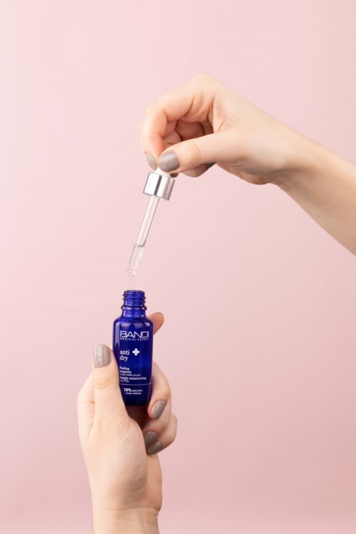 A person with painted nails uses a dropper to apply a liquid from a blue bottle with a silver cap.