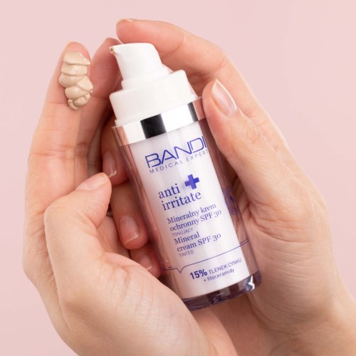 A hand holds a white bottle with a pump, labeled 'anti irritate' cream, against a light pink background.