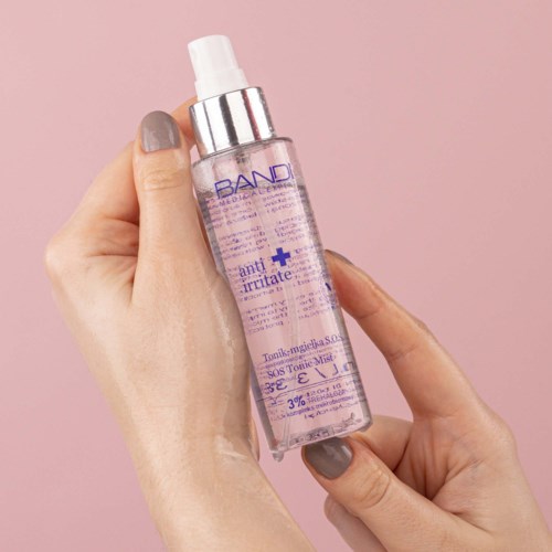 A hand holds a clear spray bottle with purple text, against a pink background.