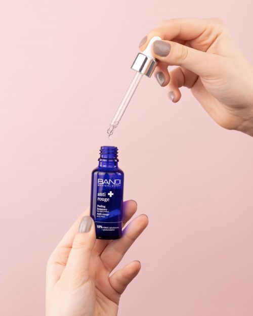 A person holds a blue bottle labeled 'BAND' and uses a dropper to add liquid. The background is light pink.