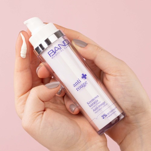 A hand holds a white and purple pump bottle labeled 'anti rouge' with cream coming out, against a pink background.