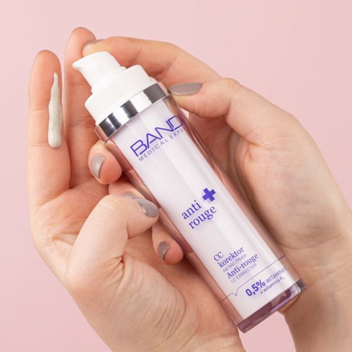 A hand holding a bottle of anti-redness cream with some cream on the finger, against a pink background.