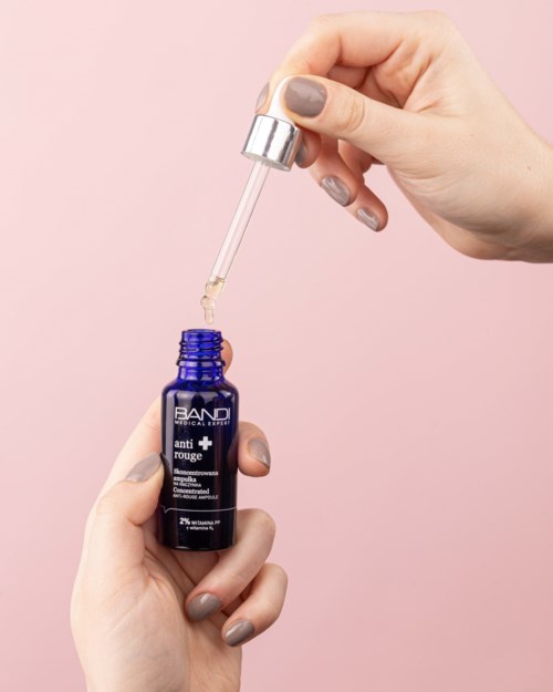 En person med malte negler bruker en dråpeteller for å påføre serum fra en blå flaske mot en rosa bakgrunn.