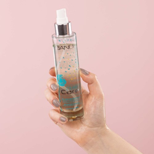 A hand holds a clear spray bottle labeled 'BAND' and 'Care' against a pink background.