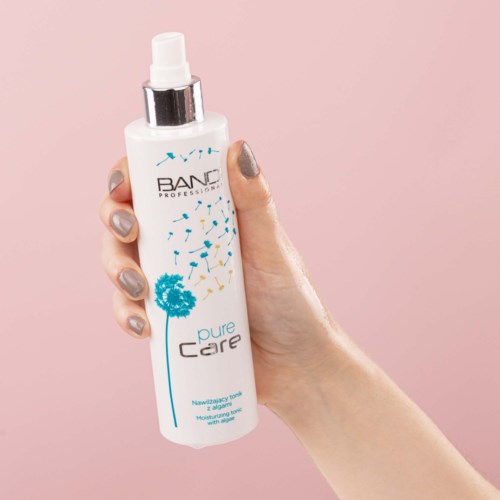 En person håller en vit sprayflaska med en maskrosdesign och etikett som säger 'Pure Care' mot en rosa bakgrund.