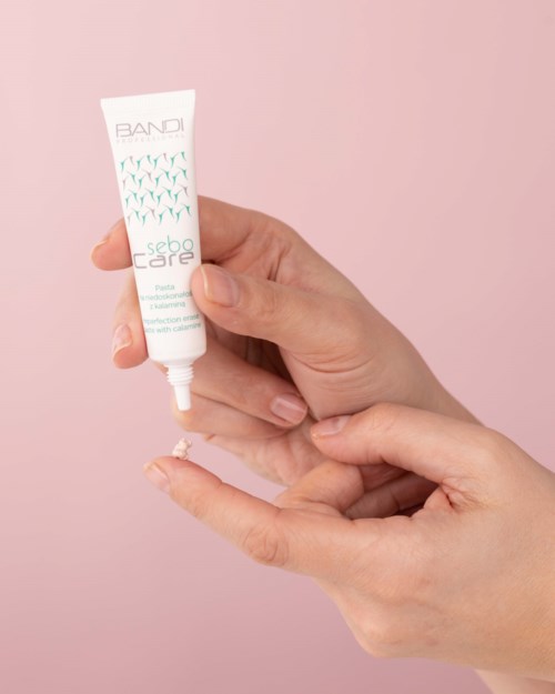 A person squeezes cream from a white tube onto their finger against a pink background.
