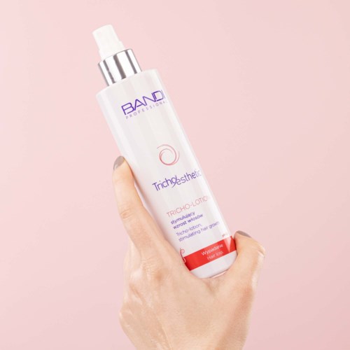 A hand holds a white bottle of skincare spray with purple and red text on a pink background.