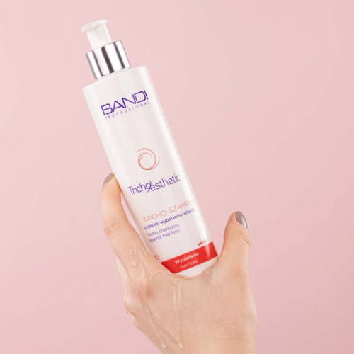 En hand håller en vit flaska med pump, märkt 'BANDI PROFESSIONAL' och 'Trichosthetic', mot en rosa bakgrund.