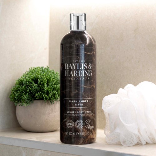 Eine dunkelbernsteinfarbene Körperwaschflasche mit silberner Kappe, neben einem weißen Waschlappen und einer grünen Pflanze auf einem Regal.