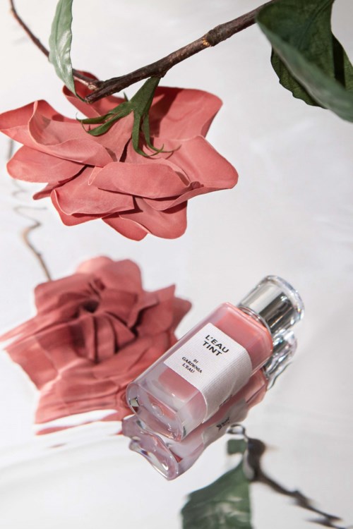 Eine rosa Nagellackflasche mit Blumenreflexion und grünen Blättern auf weißer Oberfläche.