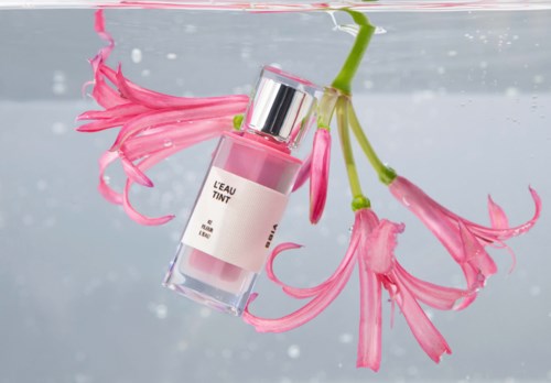 Roze bloemen drijven in water met een roze parfumfles. De fles ligt scheef en is deels onder water.